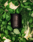Black candle jar surrounded by green basil leaves and vegetables