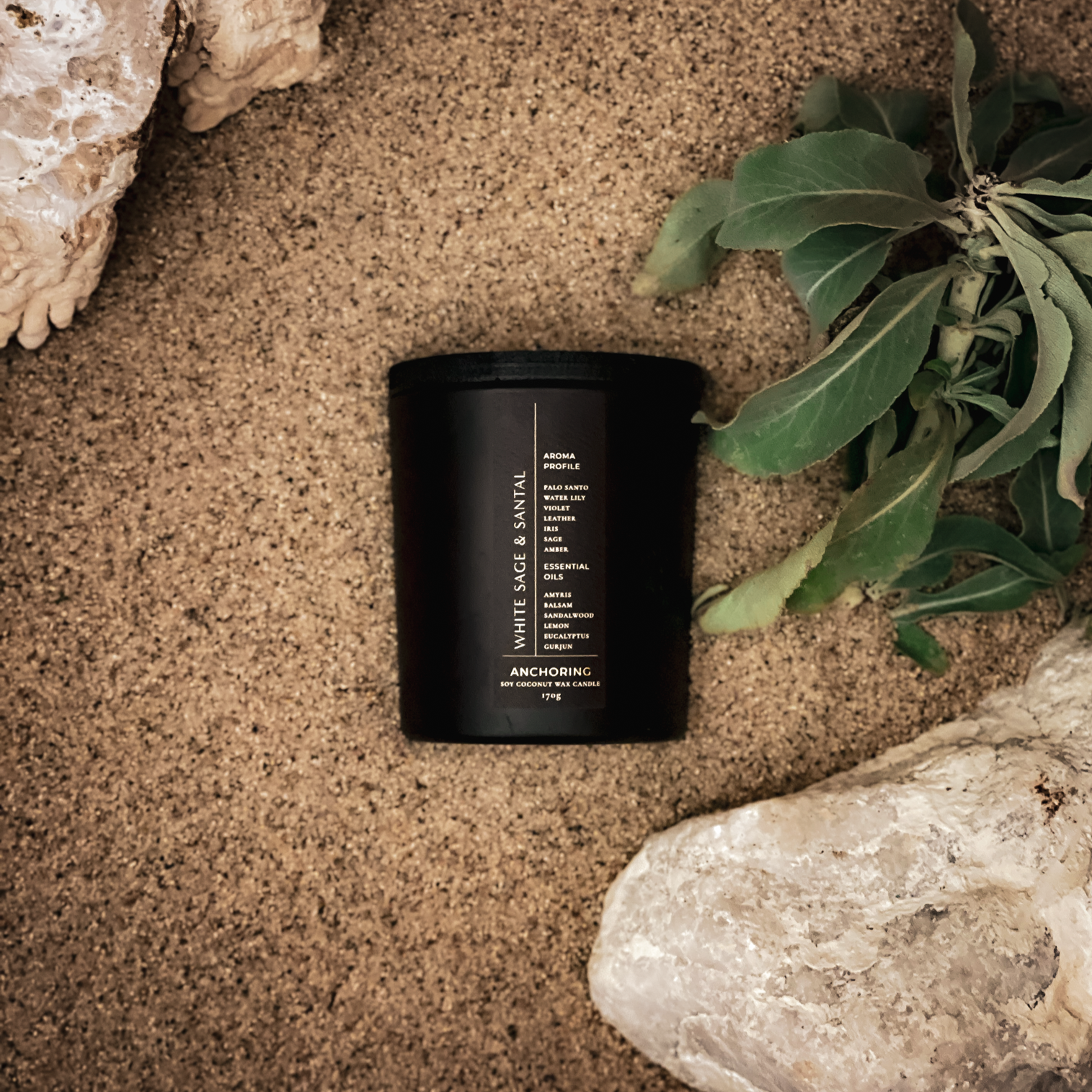 Black candle with a label on a sandy surface with rocks and plants