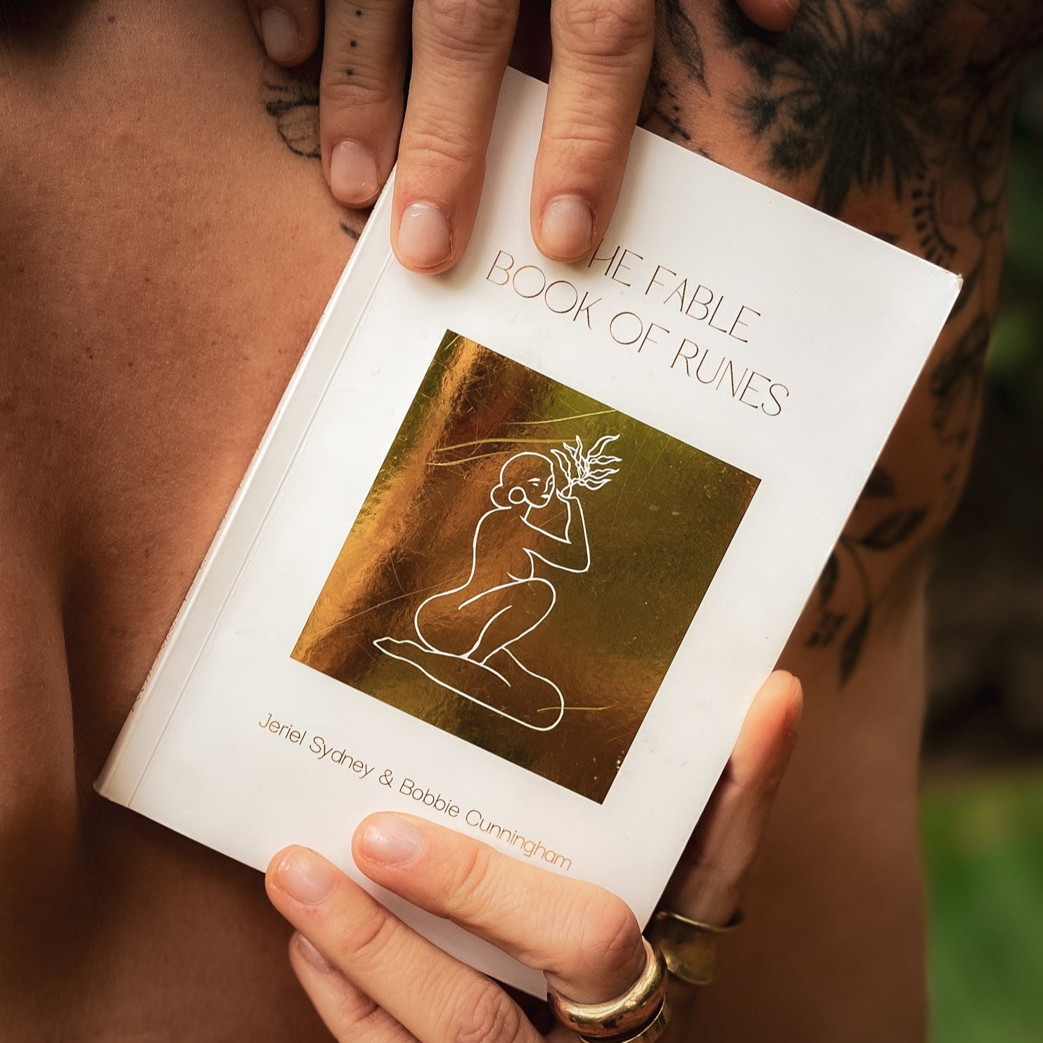 Person holding a book titled 'The Fable Book of Runes' with visible tattoos on hands.