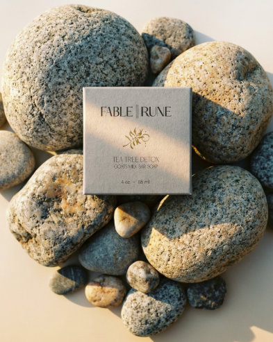 A square Fable Rune tea box placed on a bed of smooth river stones, photographed from above on a warm beige surface with soft sunlight and shadows.