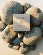 A square Fable Rune tea box placed on a bed of smooth river stones, photographed from above on a warm beige surface with soft sunlight and shadows.