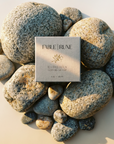 A square Fable Rune tea box placed on a bed of smooth river stones, photographed from above on a warm beige surface with soft sunlight and shadows.